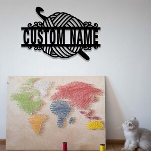 Pode incluir: Uma placa de parede de metal preto com um novelo de l&atilde; e uma agulha de croch&ecirc;, com uma barra horizontal exibindo o texto CUSTOM NAME. Abaixo, uma placa de madeira retangular apresenta um mapa-m&uacute;ndi feito com fios coloridos e pregos.
