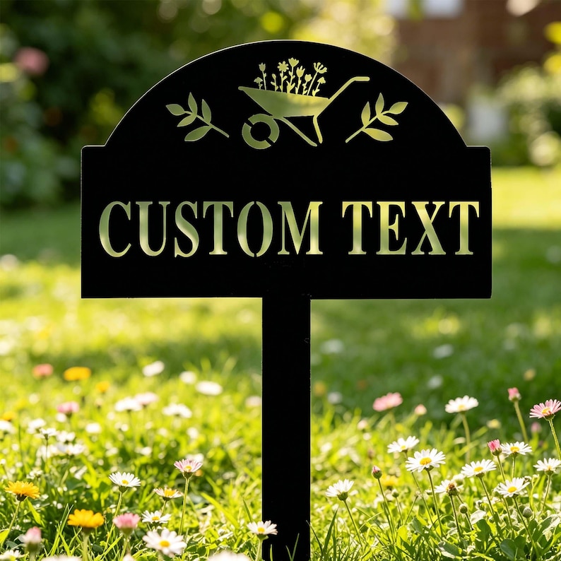 Puede incluir: Letrero de jard&iacute;n de metal negro con las palabras "CUSTOM TEXT" en dorado. La parte superior del letrero presenta un gr&aacute;fico de carretilla con flores y hojas. El letrero est&aacute; en un jard&iacute;n con flores y hierba.