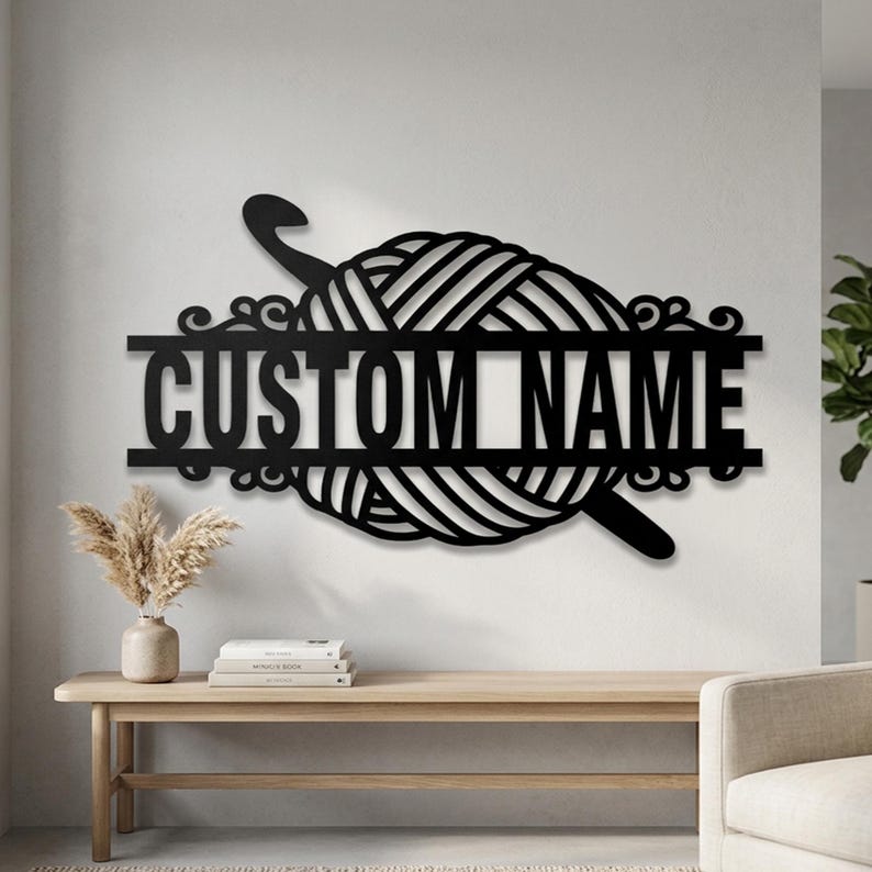 Pode incluir: Um letreiro de parede em metal preto com um novelo de l&atilde; central atravessado por uma agulha de croch&ecirc;. O design inclui uma barra horizontal no meio com o texto CUSTOM NAME em fonte sem serifa em negrito e detalhes decorativos.