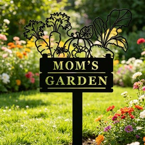 Puede incluir: Letrero de jard&iacute;n de metal negro con el texto "MOM'S GARDEN" y una parte superior decorativa con siluetas de zanahorias, una calabaza, tomates y otras verduras. El letrero est&aacute; en un jard&iacute;n con flores coloridas.