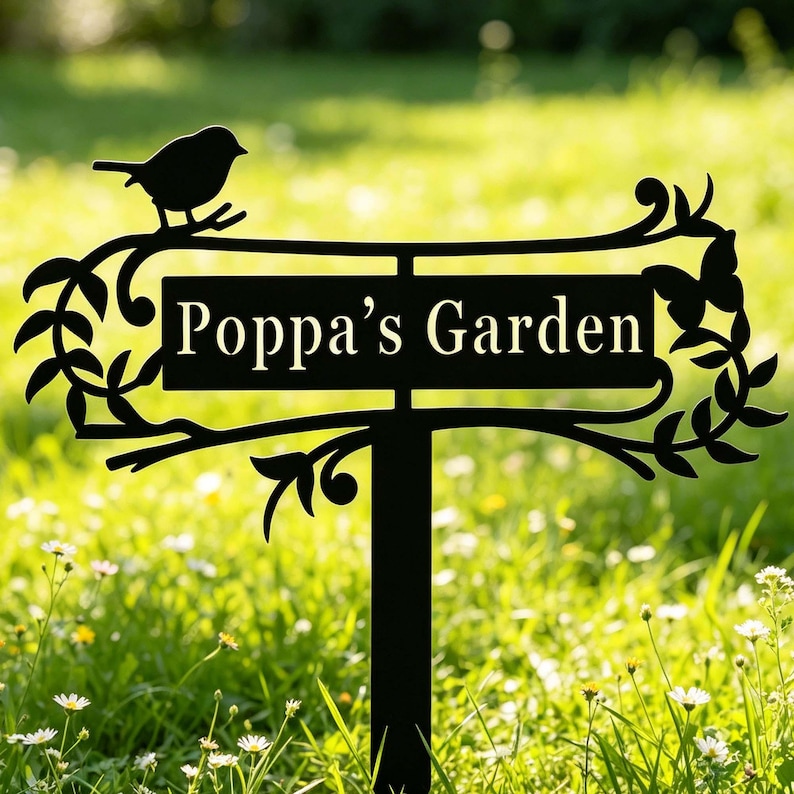 Puede incluir: Letrero de jard&iacute;n de metal negro con el texto "Poppa's Garden". El letrero presenta una silueta de un p&aacute;jaro posado en una rama, con hojas y adornos decorativos. El letrero est&aacute; montado en una estaca.