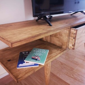 May include: A wooden TV stand with a drawer and a shelf. The stand has a light brown finish and is made of solid wood. The stand has a modern design and is perfect for any living room.