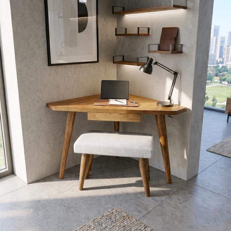 May include: A wooden corner desk with a laptop, pen, and notepad. A small, upholstered stool sits in front of the desk. A black desk lamp and floating shelves are mounted on the wall. A framed picture hangs on the wall.