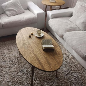 May include: A light brown oval coffee table with a wood grain finish and a book on top. The table has three legs and is sitting on a light brown shag rug.