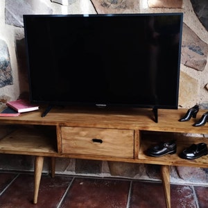 May include: A wooden TV stand with a drawer and two shelves. The stand has a dark wood finish and is sitting on a tiled floor. A flat screen TV is sitting on top of the stand. There are two pairs of shoes on the shelves.