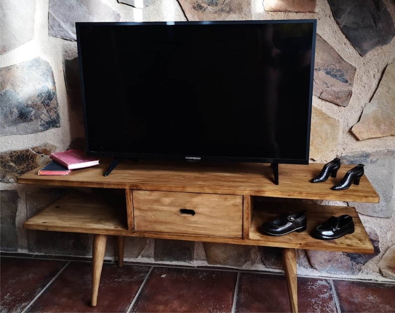 May include: A wooden TV stand with a drawer and two shelves. The stand has a dark wood finish and is sitting on a tiled floor. A flat screen TV is sitting on top of the stand. There are two pairs of shoes on the shelves.