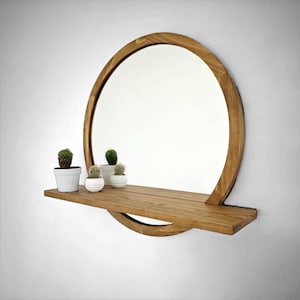 Peut inclure: Un miroir rond en bois avec une étagère, fini avec des tons de bois naturels. Le miroir a un cadre en bois épais. Trois petits cactus en pot sont exposés sur l'étagère. Le miroir est monté sur un mur blanc.