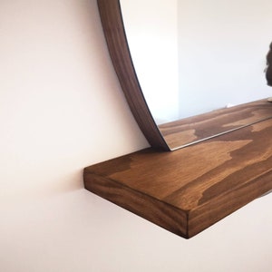 May include: A wooden shelf with a rounded mirror attached to the wall. The shelf is stained a dark brown color and has a natural wood grain pattern.