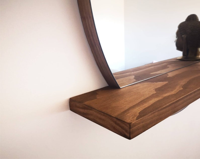 May include: A wooden shelf with a rounded mirror attached to the wall. The shelf is stained a dark brown color and has a natural wood grain pattern.