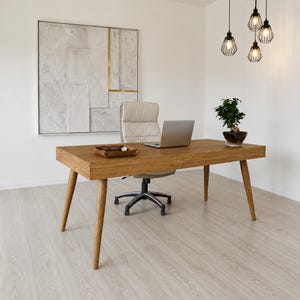 Pode incluir: Uma secretária de madeira com um laptop, uma pequena planta e uma bandeja de couro com acessórios. A secretária tem pernas anguladas e está numa sala bem iluminada com uma pintura moderna e luzes penduradas. A secretária tem cerca de 152 cm de comprimento.