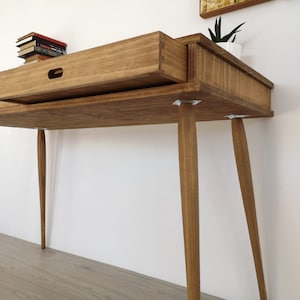 May include: A wooden desk with two drawers and tapered legs. The desk is made of light brown wood and has a natural finish.