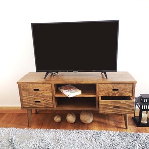 May include: A wooden TV stand with two drawers and a shelf. The stand has a dark brown finish and is sitting on a light brown wood floor. There is a flat screen TV on top of the stand and a book on the shelf. There are two round, light brown balls on the floor in front of the stand.
