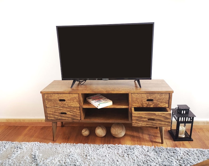 May include: A wooden TV stand with two drawers and a shelf. The stand has a dark brown finish and is sitting on a light brown wood floor. There is a flat screen TV on top of the stand and a book on the shelf. There are two round, light brown balls on the floor in front of the stand.
