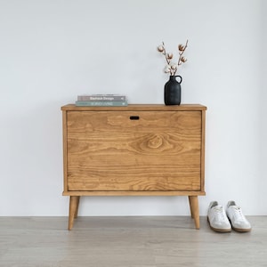May include: A light brown wooden cabinet with a single door and four angled legs. The cabinet has a small handle and is topped with books and a black vase with cotton stems. White trainers are placed on the floor in front of the cabinet.