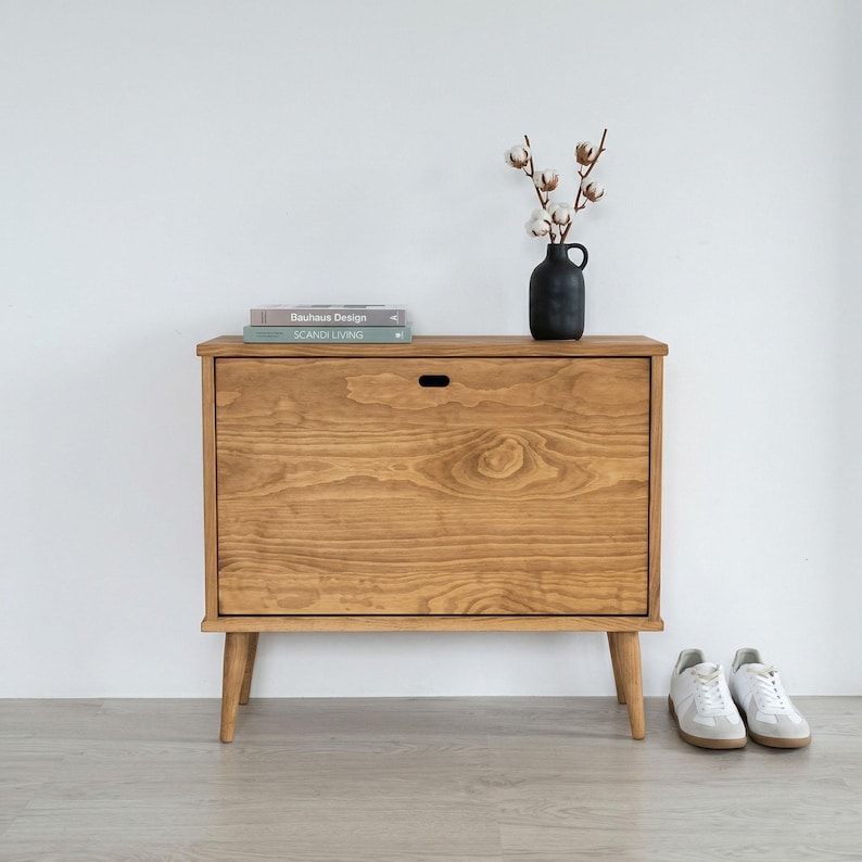 May include: A light brown wooden cabinet with a single door and four angled legs. The cabinet has a small handle and is topped with books and a black vase with cotton stems. White trainers are placed on the floor in front of the cabinet.