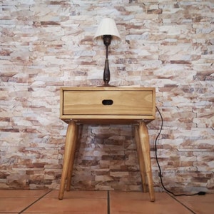 Op de afbeelding: Een houten nachtkastje met een lade en taps toelopende poten. Een witte lampenkap met een zwarte lampvoet staat op het nachtkastje.