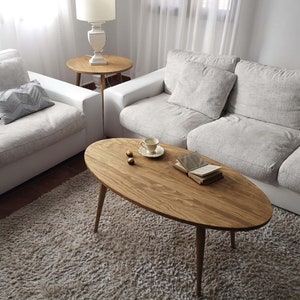 May include: A light brown oval coffee table with three legs sits on a fluffy white rug in a living room. The table has a natural wood grain finish and is surrounded by white couches.