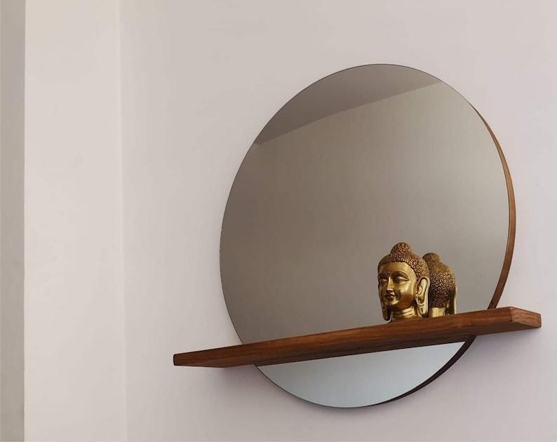 May include: A round mirror with a wooden shelf underneath. Two gold statues of Buddha are on the shelf.