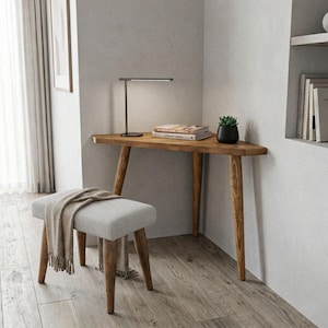 Könnte beinhalten: Ein Holztisch mit dreieckiger Platte und angewinkelten Beinen, kombiniert mit einem grau gepolsterten Hocker. Eine schwarze Schreibtischlampe, eine kleine Topfpflanze und Bücher stehen auf dem Tisch. Eine Fransen-Decke liegt auf dem Hocker.