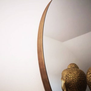 May include: A round mirror with a wooden frame. The frame is a light brown color and has a natural wood grain. The mirror is reflecting two gold statues of Buddha.
