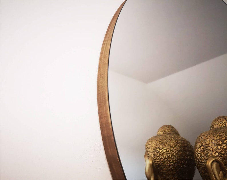 May include: A round mirror with a wooden frame. The frame is a light brown color and has a natural wood grain. The mirror is reflecting two gold statues of Buddha.