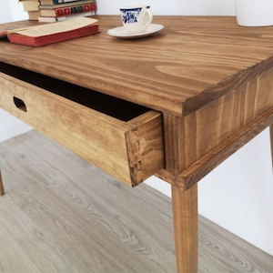 May include: A wooden desk with a single drawer and tapered legs. The desk is stained a light brown color and has a natural wood grain finish.
