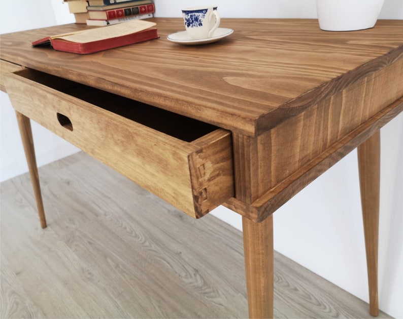 May include: A wooden desk with a single drawer and tapered legs. The desk is stained a light brown color and has a natural wood grain finish.