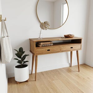 Può includere: Una consolle in legno in stile moderno di metà secolo, con due cassetti e un ripiano. Uno specchio rotondo dorato è appeso sopra. Il tavolo ha gambe affusolate e una finitura in legno naturale. Una borsa tote è appesa al muro.