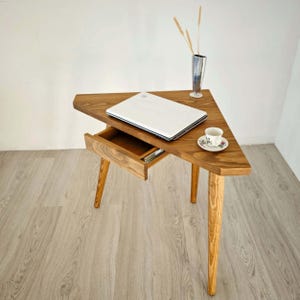Könnte beinhalten: Ein dreieckiger Holzschreibtisch mit einer Schublade und drei Beinen. Ein Laptop steht auf dem Schreibtisch, und eine kleine Tasse und Untertasse befinden sich auf der rechten Seite des Schreibtisches.