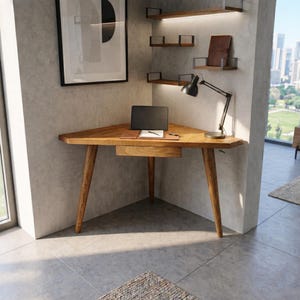 May include: A wooden corner desk with a triangular top and tapered legs. A laptop, notepad, and pen sit on the desk. A black desk lamp is positioned on the desk. The desk is in a modern office setting.