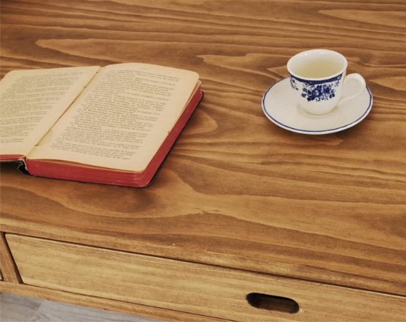 May include: A wooden table with a drawer and a book lying open on the surface. A cup and saucer with a blue and white floral pattern are on the table.