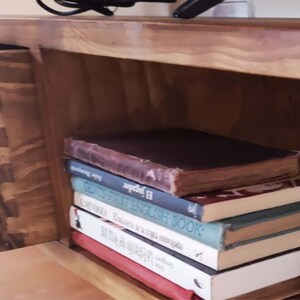 May include: A wooden shelf unit with a drawer and a stack of books. The top book is brown with a gold spine and the text "El Indigena" is visible. The other books are various colours and have titles like "First English Book" and "The History of a Cat."