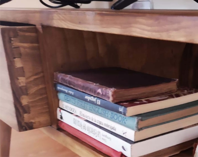 May include: A wooden shelf unit with a drawer and a stack of books. The top book is brown with a gold spine and the text "El Indigena" is visible. The other books are various colours and have titles like "First English Book" and "The History of a Cat."