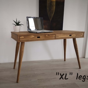 May include: A wooden desk with two drawers and long legs. The desk is made of natural wood and has a light brown finish. The desk has a laptop on top of it and a small plant in a white pot. The text "XL" legs is written on the floor in front of the desk.