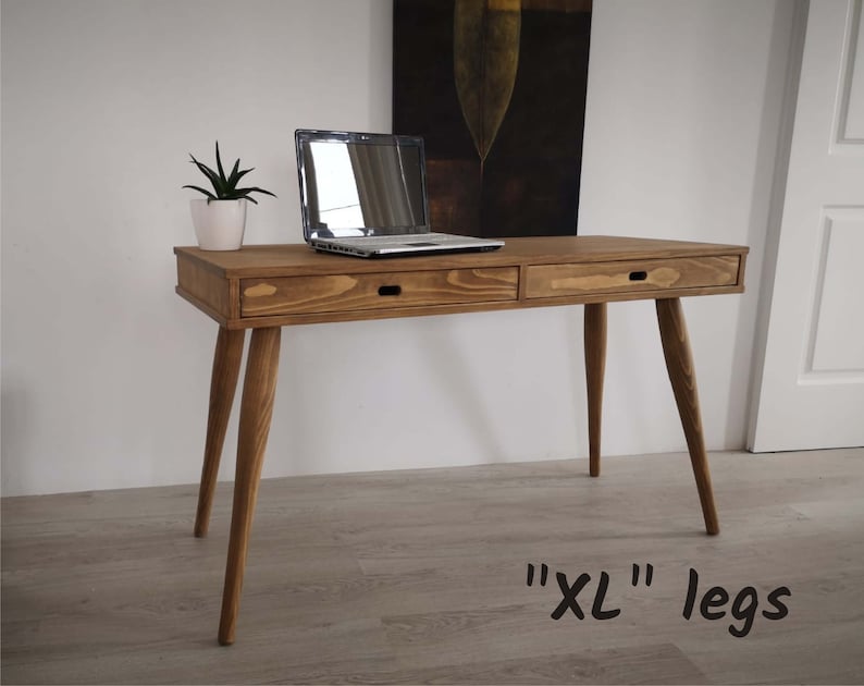 May include: A wooden desk with two drawers and long legs. The desk is made of natural wood and has a light brown finish. The desk has a laptop on top of it and a small plant in a white pot. The text "XL" legs is written on the floor in front of the desk.