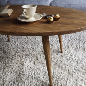 May include: A wooden oval coffee table with four tapered legs. The table is sitting on a fluffy white rug. A white teacup and saucer and a small bowl of wrapped chocolates are on the table.