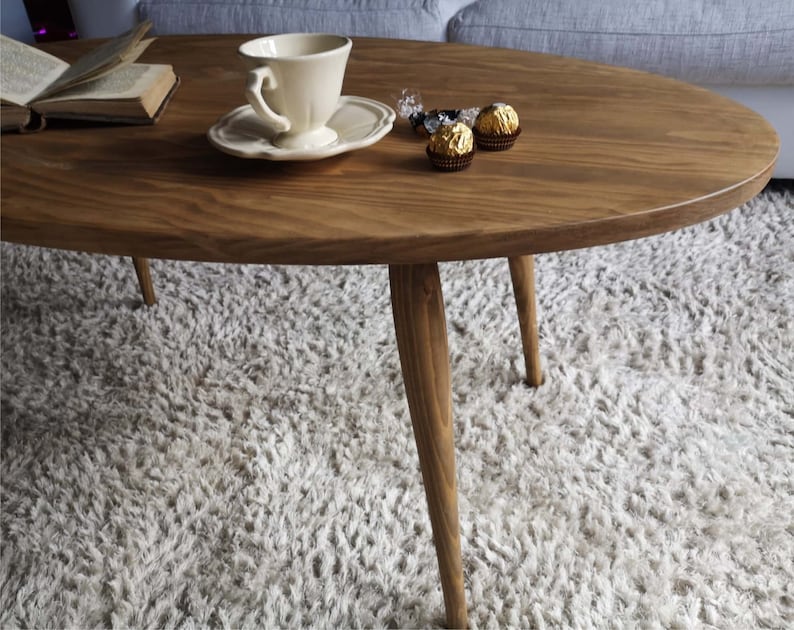 May include: A wooden oval coffee table with four tapered legs. The table is sitting on a fluffy white rug. A white teacup and saucer and a small bowl of wrapped chocolates are on the table.