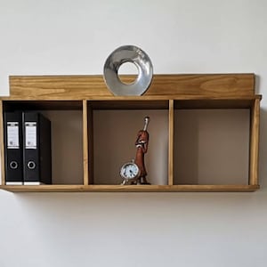 Op de afbeelding: Een houten wandplank met drie compartimenten. Twee zwarte ordners staan in het linkervak. Een zilveren metalen ring en een klein beeldje met een klok staan op de plank.