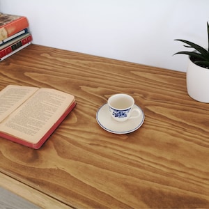 May include: A wooden desk with a book, a cup and saucer, and a potted plant. The desk has a natural wood finish and is made of solid wood.