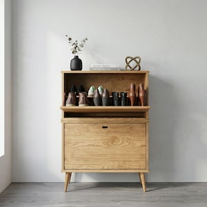 May include: A light-coloured wooden shoe cabinet with a top shelf holding various shoes. The cabinet has a drawer and a slatted top section. A black vase with greenery and a decorative knot sit on top.