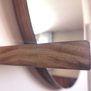 May include: A close-up of a wooden shelf attached to a round mirror. The shelf is made of light brown wood with a natural grain pattern.