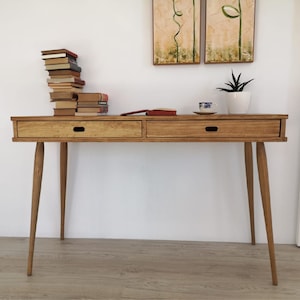 May include: A light wood desk with two drawers and tapered legs. The desk has a natural wood finish and is sitting on a wood floor. There are books stacked on the desk, a cup of tea, and a potted plant.