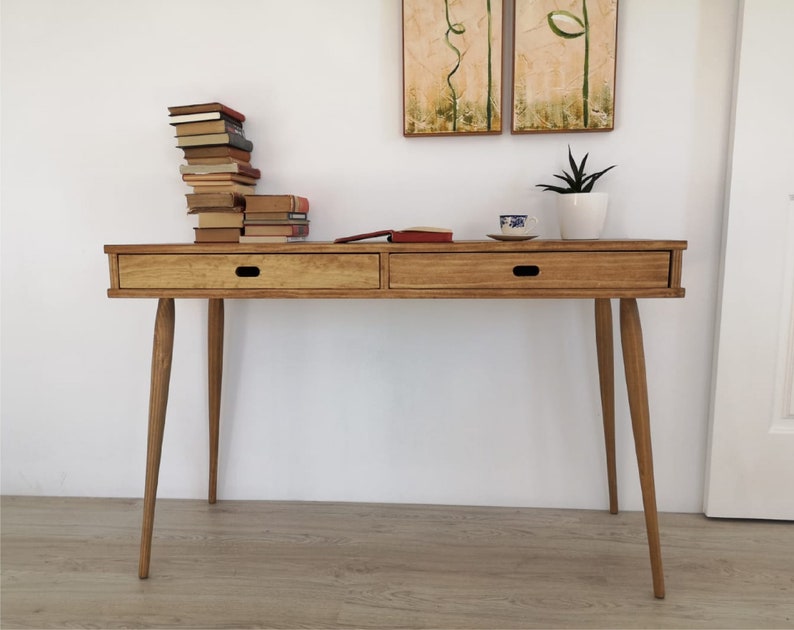 May include: A light wood desk with two drawers and tapered legs. The desk has a natural wood finish and is sitting on a wood floor. There are books stacked on the desk, a cup of tea, and a potted plant.
