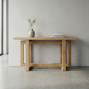 May include: A light-coloured wooden console table with a rectangular top and a lower shelf. The table has a sturdy, geometric design. A small, light green vase with dried plants and a stack of books are on the top. The table is approximately 152 cm long.