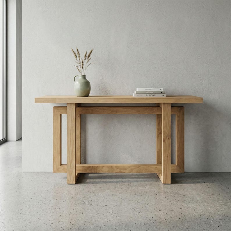 May include: A light-coloured wooden console table with a rectangular top and a lower shelf. The table has a sturdy, geometric design. A small, light green vase with dried plants and a stack of books are on the top. The table is approximately 152 cm long.