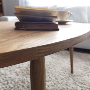 May include: A light brown oval coffee table with three legs. The table is made of wood and has a smooth, polished finish.