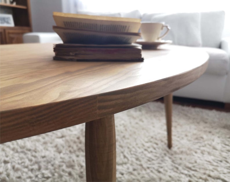May include: A light brown oval coffee table with three legs. The table is made of wood and has a smooth, polished finish.
