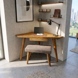 May include: A wooden corner desk with a small drawer, a laptop, and a black desk lamp. A small upholstered stool sits in front of the desk. The desk is set in a corner with a window and shelves in the background.