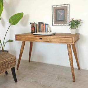 Peut inclure: Un bureau en bois brun clair avec un seul tiroir et des pieds effilés. Le bureau a un ordinateur portable, une petite figurine de chien en céramique et une pile de livres dessus.