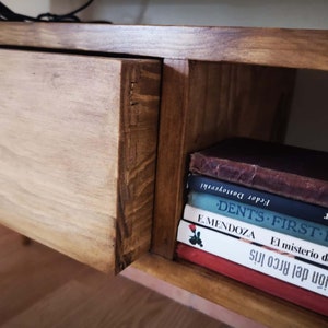 May include: A wooden shelf unit with a drawer and a shelf. The shelf is filled with books, including "DENT'S FIRST ENGLISH" and "El misterio de la cript."